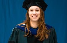 Woman wearing academic regalia smiling in front of a blue backdrop