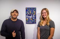 Two students smiling and standing against a wall next to a poster