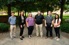 6 people stand facing the camera standing on a sidewalk with trees behind them