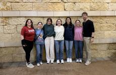 Seven people dressed nicely and standing on a sidewalk with a stone wall or building behind them