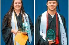 two students wearing graduation attire and holding glass awards