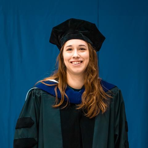 Woman wearing academic regalia smiling in front of a blue backdrop