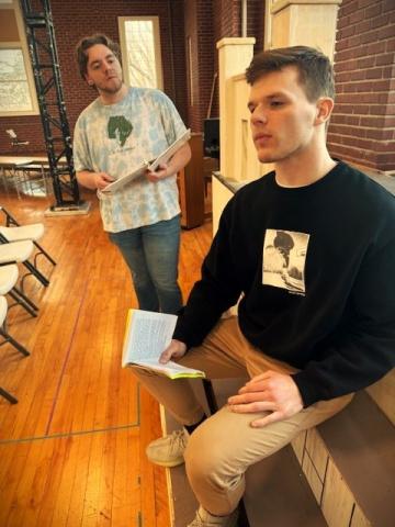 Two actors rehearsing theatre play, one is holding a book looking into the distance as the other holds a book and looks at the first actor