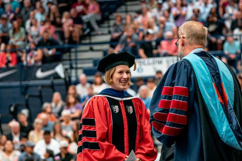Woman wearing academic regalia smiling in front of crowd Woman wearing academic regalia smiling in front of crowd