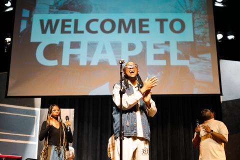 Sterling College Chapel featuring man singing and clapping his hands on stage with a Screen behind him