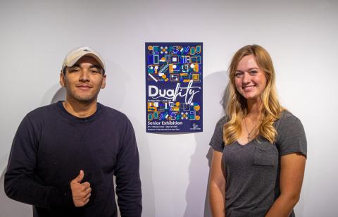 Two students smiling and standing against a wall next to a poster Two students smiling and standing against a wall next to a poster