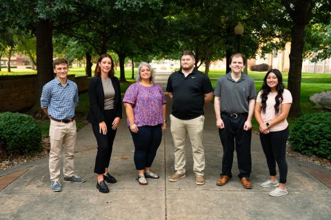 6 people stand facing the camera standing on a sidewalk with trees behind them