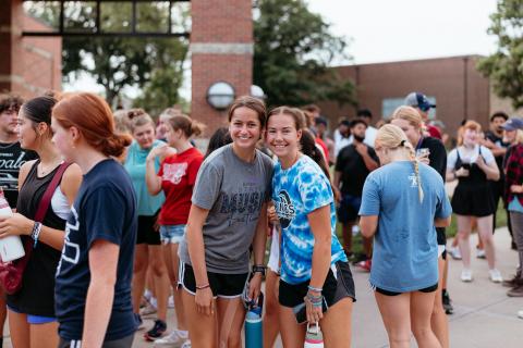 Two girls stand in the middle of a crowd of people, smiling at the camera