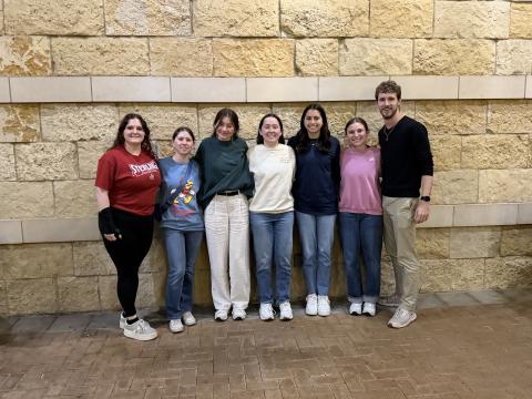 Seven people dressed nicely and standing on a sidewalk with a stone wall or building behind them