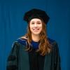 Woman wearing academic regalia smiling in front of a blue backdrop