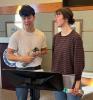 students rehearsing for music, both holding objects, one looking at the other and one looking at a music stand