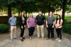 6 people stand facing the camera standing on a sidewalk with trees behind them